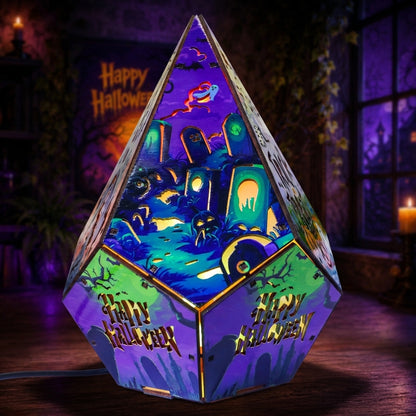 A geometric, pyramid-shaped Halloween lamp with a multicolor printed design featuring tombstones, bats, and the words 'Happy Halloween' on the sides, shown lit in a spooky room setting.